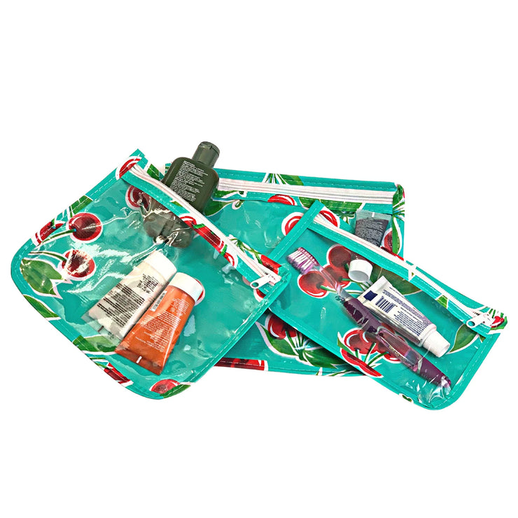 Above view: Small, medium and large slim travel bags, white zippers and cherry print on green background, showing travel items through transparent front. 