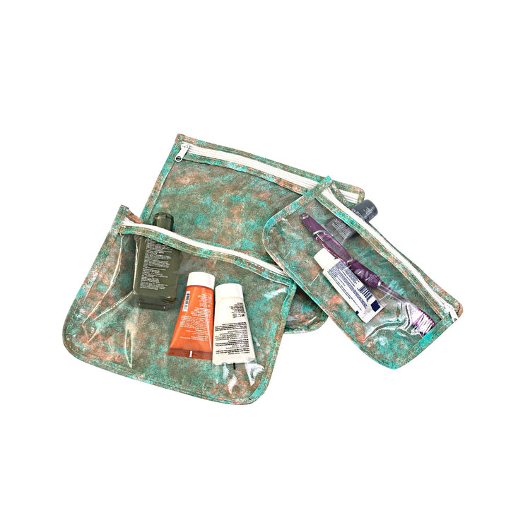 Above view: Small, medium and large slim travel bags, white zippers and green, orange, olive splatter print, showing travel items through transparent front. 