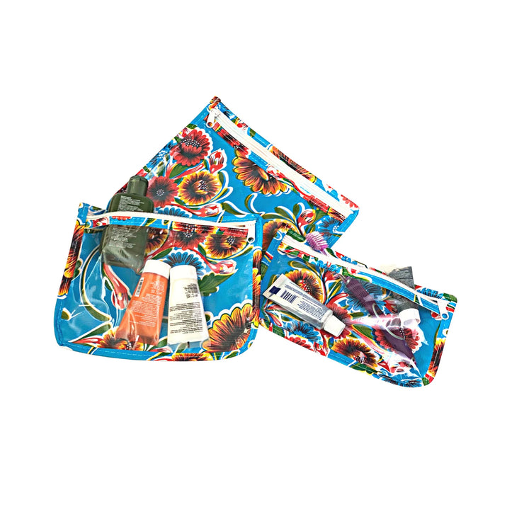 Above view: Small, medium and large slim travel bags, white zippers and multi-colored floral print on blue background, showing travel items through transparent front.