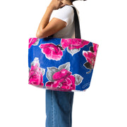 Front view: Woman models large pink rose print on blue background tote with black handles.