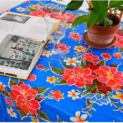 Above view: Multi-colored hibiscus print on blue background tablecloth covering table, open book displayed and potted plant centerpiece.