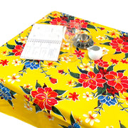 Above view: Multi-colored hibiscus print on yellow background tablecloth covering table, open notebook displayed.