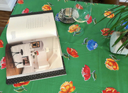 Above view: Multi-colored flower print on green background tablecloth, open book displayed with potted plant centerpiece. 